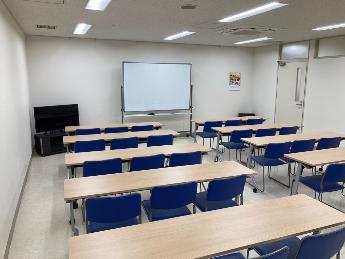 会議室2。机と青い椅子、スクリーンが見える。